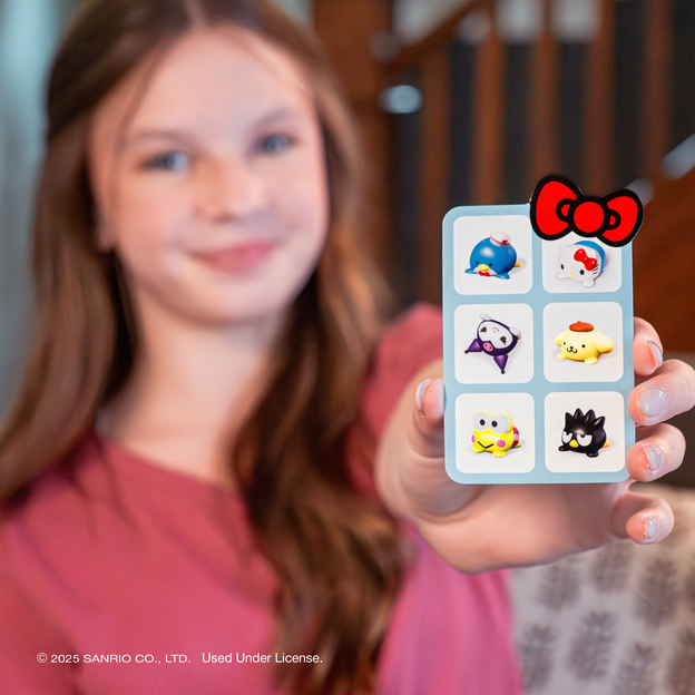 A teenage girl with long brunette hair, wearing a pink t-shirt is holding out a card with 6 assorted characters from the Hello Kitty and Friends Reach For the Stars Game.