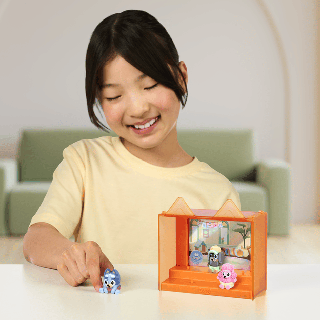 Young girl sitting down, smiling, playing with 3 fuzzy Bluey characters. She's holding one figure in her hand, the other 2 are sitting in their display case. 