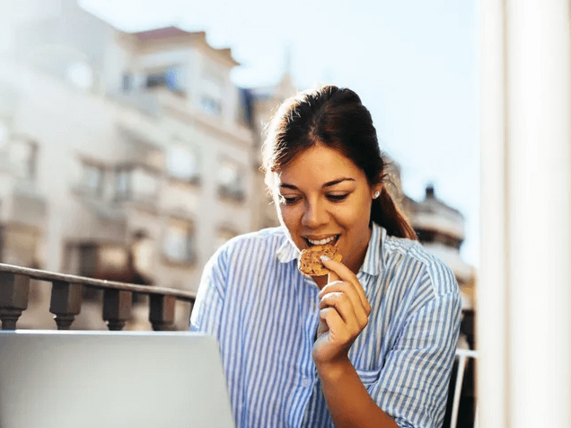 Joven realizando test de inglés online con Berlitz en su laptop desde una terraza, comiendo una galleta