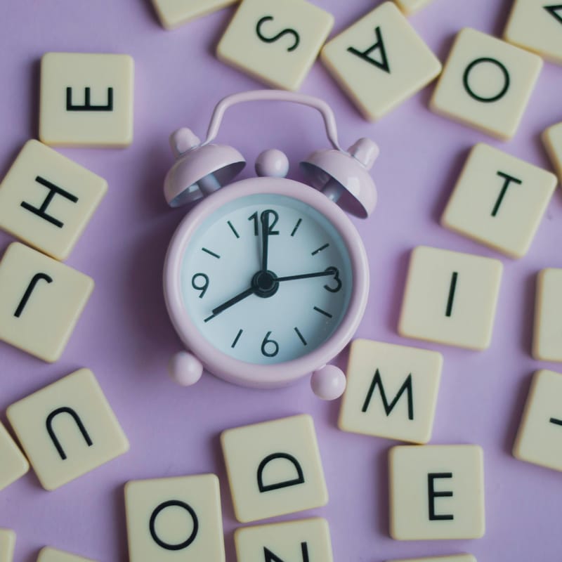 Reloj rosa pastel rodeado de letras en fondo morado pastel para representar la frecuencia en inglés.