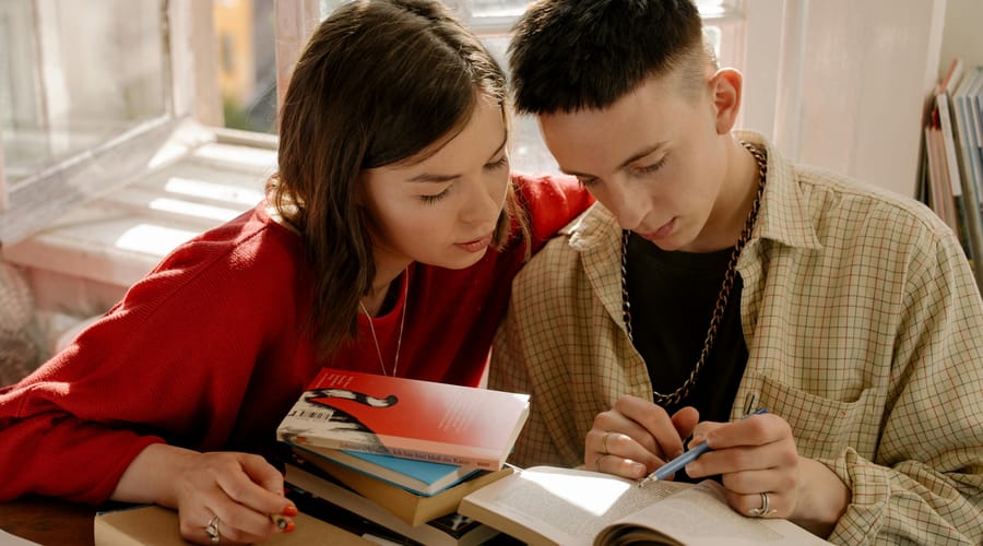 Dos jóvenes sentados leyendo un libro juntos, acompañados de varios libros sobre cuál es el idioma más antiguo del mundo.